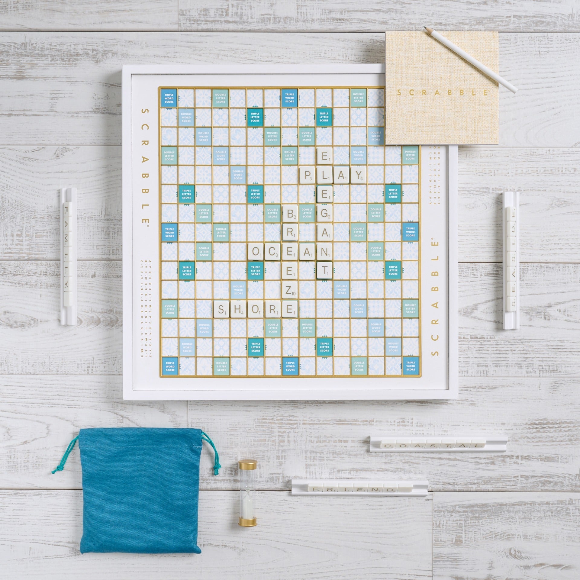The Scrabble Bianco Edition by WS Game Company features a wood-framed board with tiles spelling "PLAY," "BIG," "OCEAN," "BREEZE," "SHORE," and "ELEGANT." A blue tile bag, score pads, and sand timer sit beside it on a wooden surface, perfect for an ocean breeze design.