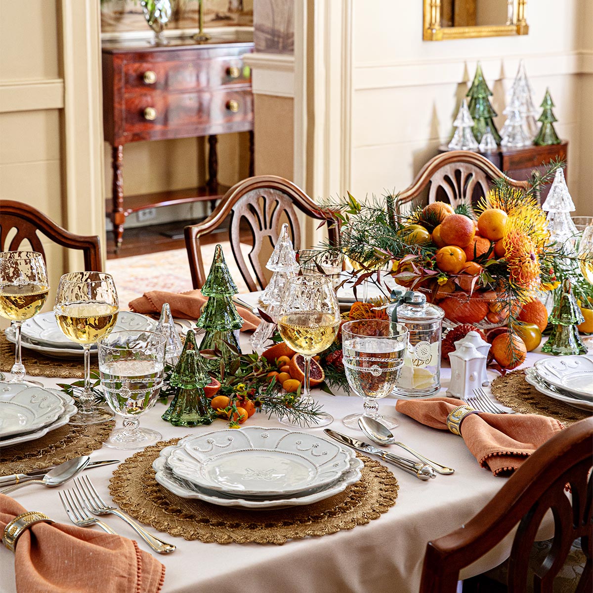 A dining table set for four with white plates, wine glasses, woven placemats, orange napkins, Berry & Thread details by Juliska, vibrant fruit centerpiece, and Juliska’s Berry & Thread Evergreen Tree Collection as festive holiday decor.
