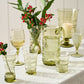 A curated selection from Juliska’s Ella Drinkware Collection—featuring vibrant green glasses, a pitcher, a vase filled with red berries, and an elegant candle—each piece beautifully crafted from mouth-blown glassware and artfully arranged atop a crisp white tablecloth.