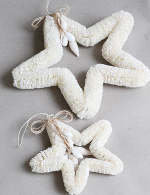 The Dekorasyon Bottlebrush and Shell Star Ornament features two white star-shaped designs crafted from textured abaca fiber, adorned with twine bows and decorative white shells for a charming coastal touch.