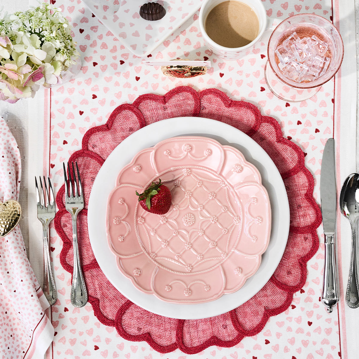 A pink plate with a strawberry sits atop the Heart Placemat Collection by Juliska, surrounded by cutlery, a coffee cup, glass of water, and flowers on an elegant tablescape adorned with embroidered hearts.