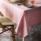 A Juliska Pink Berry Scallop Tablecloth adorns the table, set with plates, cutlery, a pink glass, and accompanied by a woven chair.