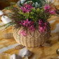 The Juliska Provence Rattan Turtle Bowl, hand-woven and filled with pink and green flowers, rests on a patterned tablecloth with other tableware, bringing natural texture and charm to the setting.