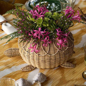 The Juliska Provence Rattan Turtle Bowl, with its natural texture, holds green and pink flowers and is placed on a yellow and white tablecloth.