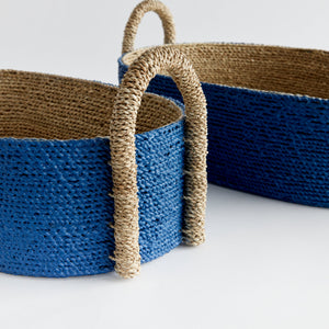 The Tiana Blue Seagrass Short Oval Basket Collection by Napa Home & Garden features two handwoven baskets with blue bases and natural handles, ideal for enhancing your space with stylish designer home decor.