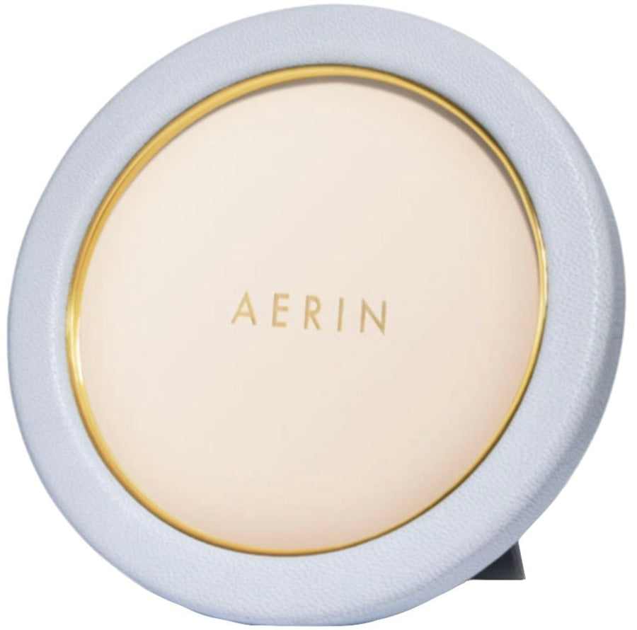The Piero Leather Frame Collection by Aerin features a round tabletop mirror with a light blue frame, Italian leather accents, and gold detailing for an elegant display.