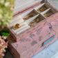 The Addison Ross Pink Chinoiserie Box Collection, a high gloss pink floral jewelry box, organizes rings, necklaces, and earrings on a marble surface with blurred flowers nearby.