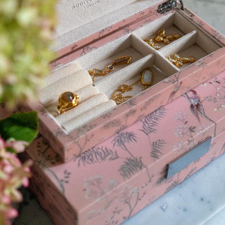 The Addison Ross Pink Chinoiserie Box Collection, a high gloss pink floral jewelry box, organizes rings, necklaces, and earrings on a marble surface with blurred flowers nearby.