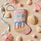 The Palm Beach Holiday Needle Point Stocking by Bauble Stockings, featuring an umbrella and chairs design, is displayed on sand surrounded by seashells, pastel ribbons, and paper drink umbrellas.