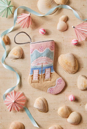 The Palm Beach Holiday Needle Point Stocking by Bauble Stockings, featuring an umbrella and chairs design, is displayed on sand surrounded by seashells, pastel ribbons, and paper drink umbrellas.