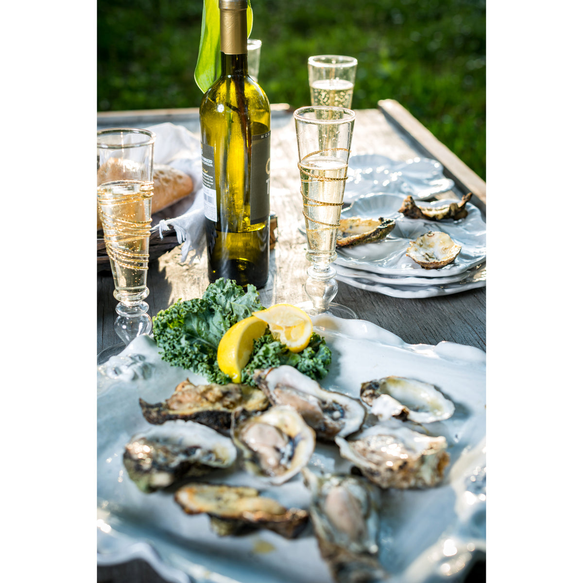 A sunny outdoor table features the Abigails Fleur De Lis Oyster Plate with Oysters Rockefeller and lemon garnish, paired with a bottle of white wine and filled glasses.