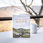 The 100-day devotional "Resilient Hope" by Thomas Nelson/Harper Collins sits on a table next to a white mug, with softly blurred mountains and trees in the background.