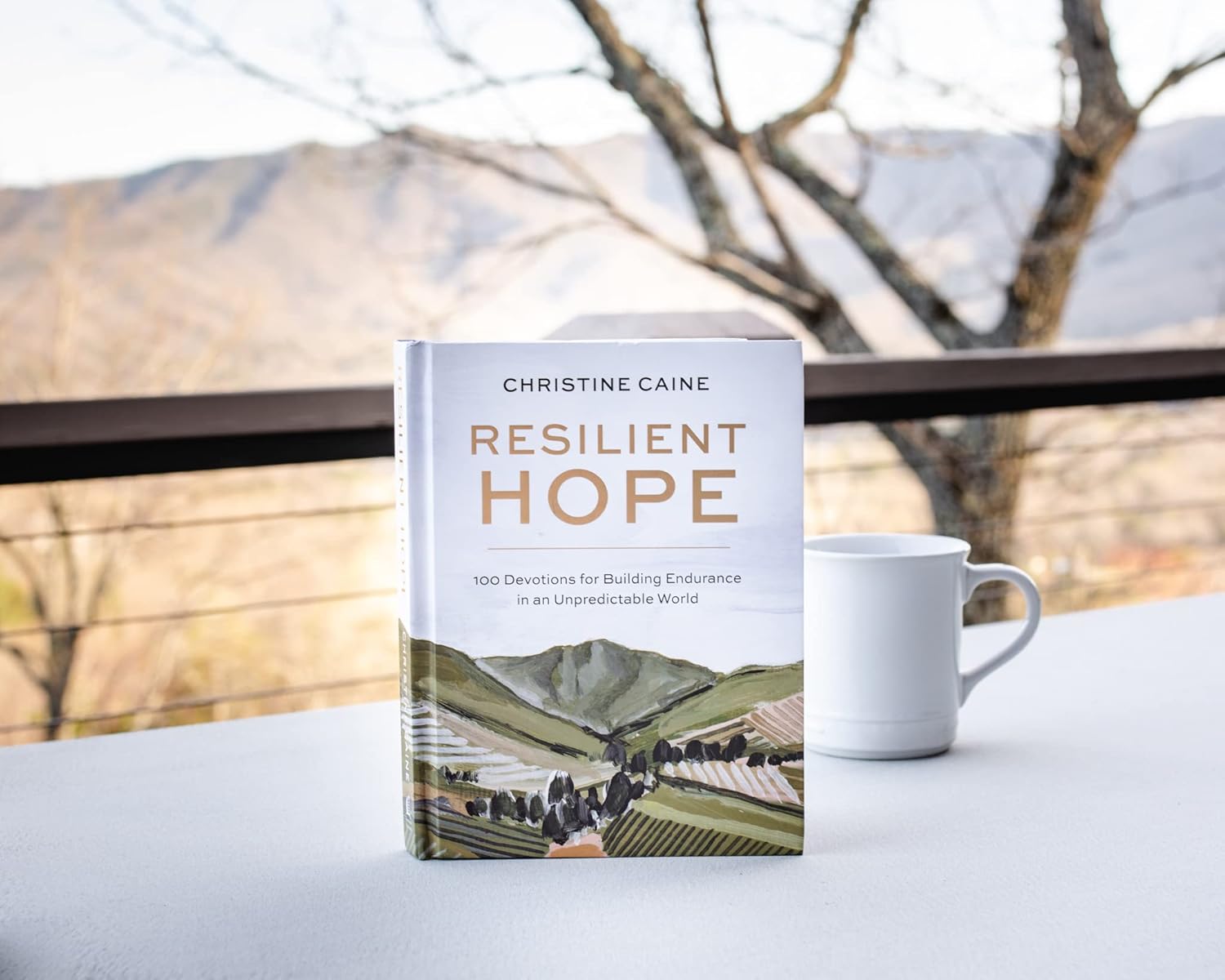 The 100-day devotional "Resilient Hope" by Thomas Nelson/Harper Collins sits on a table next to a white mug, with softly blurred mountains and trees in the background.