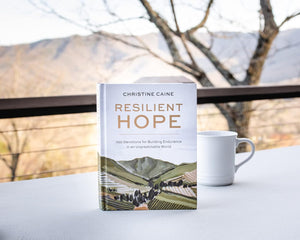 The 100-day devotional "Resilient Hope" by Thomas Nelson/Harper Collins sits on a table next to a white mug, with softly blurred mountains and trees in the background.