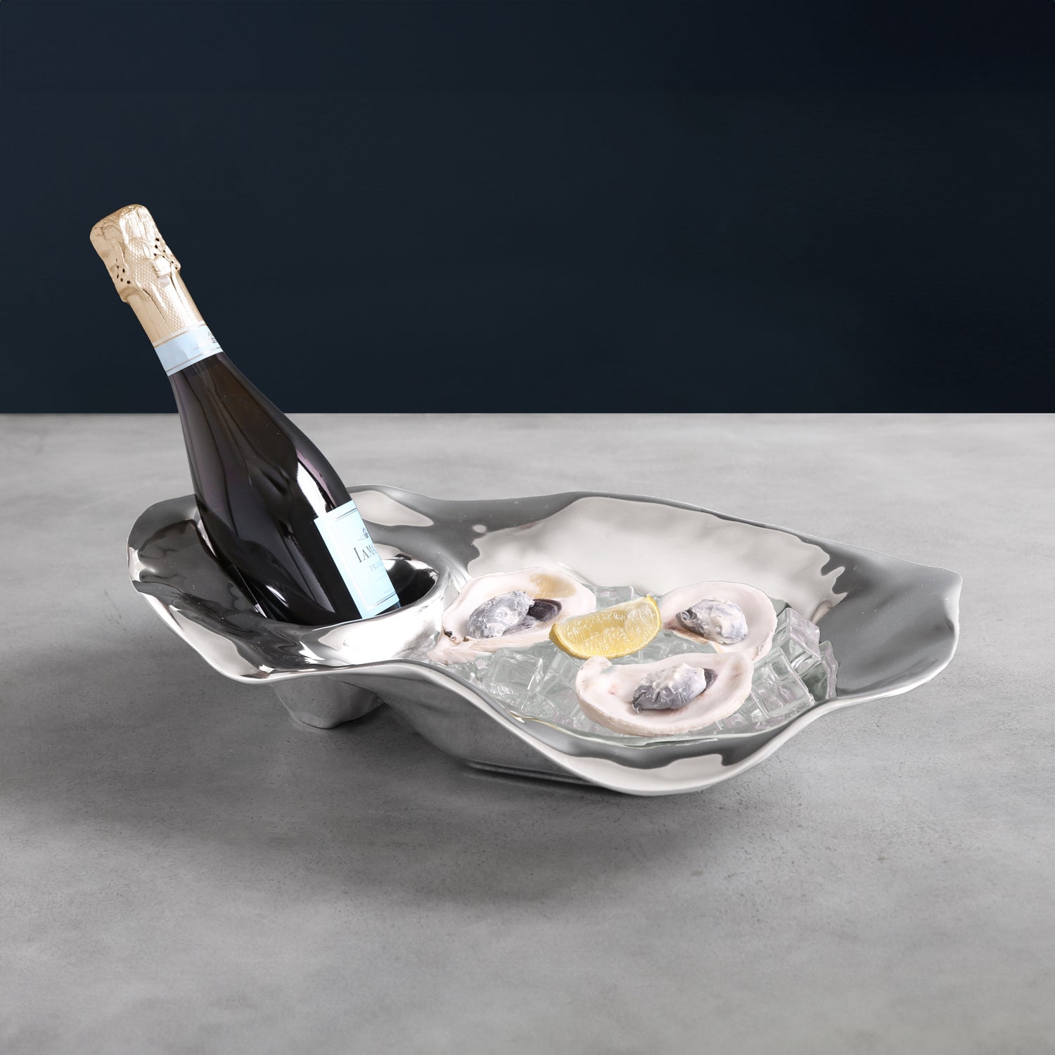 The Beatriz Ball Ocean Oyster Champagne Bucket Collection elegantly displays a bottle of champagne and three oysters on ice with a lemon wedge, all arranged on a gray surface.