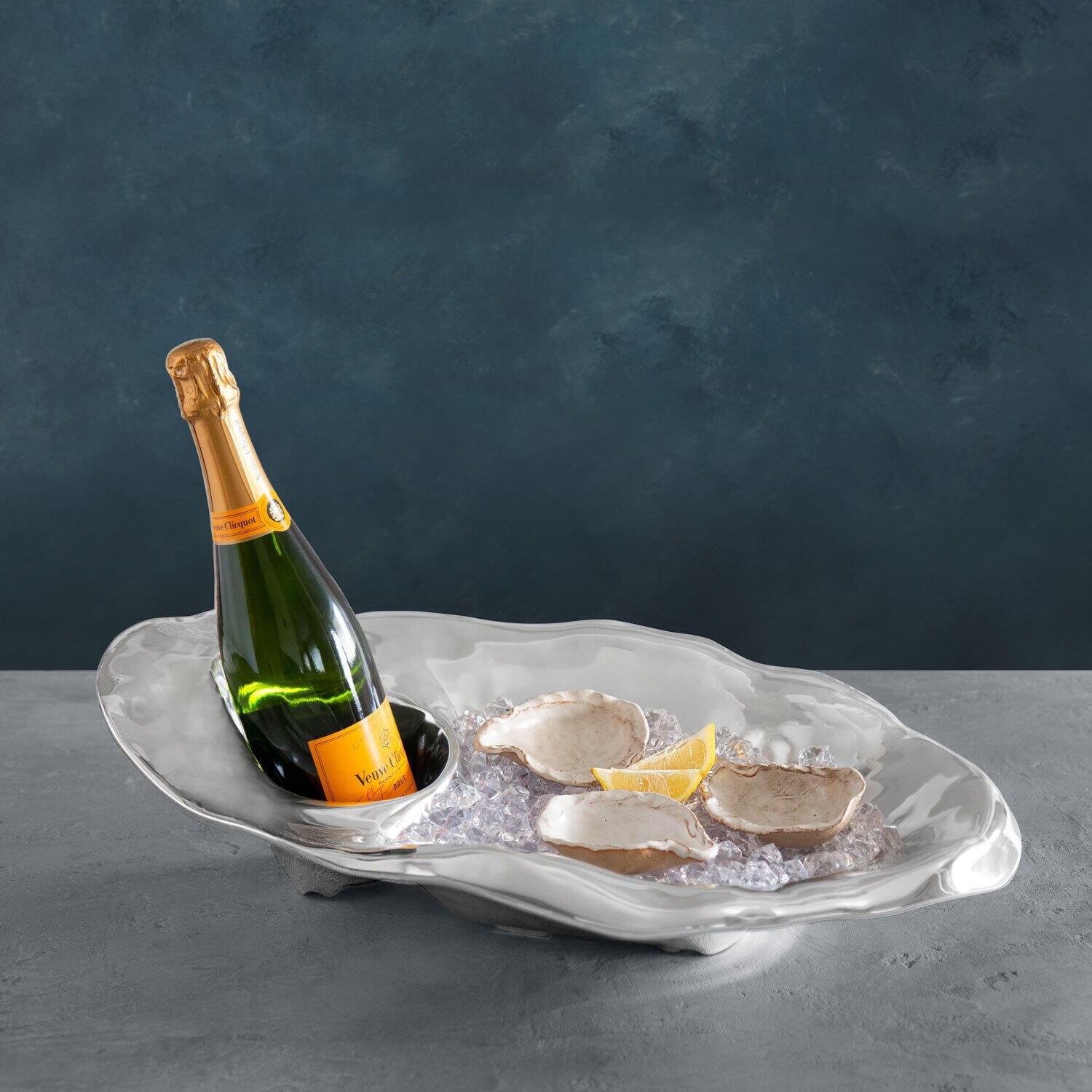 A bottle of champagne and three oysters with lemon wedges rest on ice in a clam-shaped Beatriz Ball Ocean Oyster Champagne Bucket Collection serving dish.
