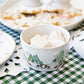 A Juliska Berry & Thread North Pole bowl, featuring a winter scene, holds large marshmallows atop a green checkered tablecloth next to a plate of cookies.