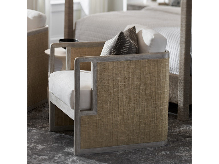 The PALECEK Aveline Lounge Chair features a hardwood frame, beige woven sides, and white cushions, shown on a patterned rug beside a bed with neutral bedding.