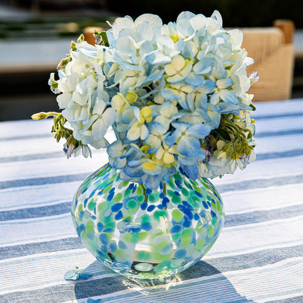 The Juliska Puro Speckled Vase by Juliska, featuring blue-green speckles, holds pale blue hydrangeas and sits atop a striped tablecloth.