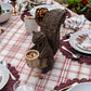 The Juliska Devon Willow Squirrel Flameless Candle Holder serves as a charming centerpiece on a plaid tablecloth, surrounded by plates, glasses, and utensils on the dining table.