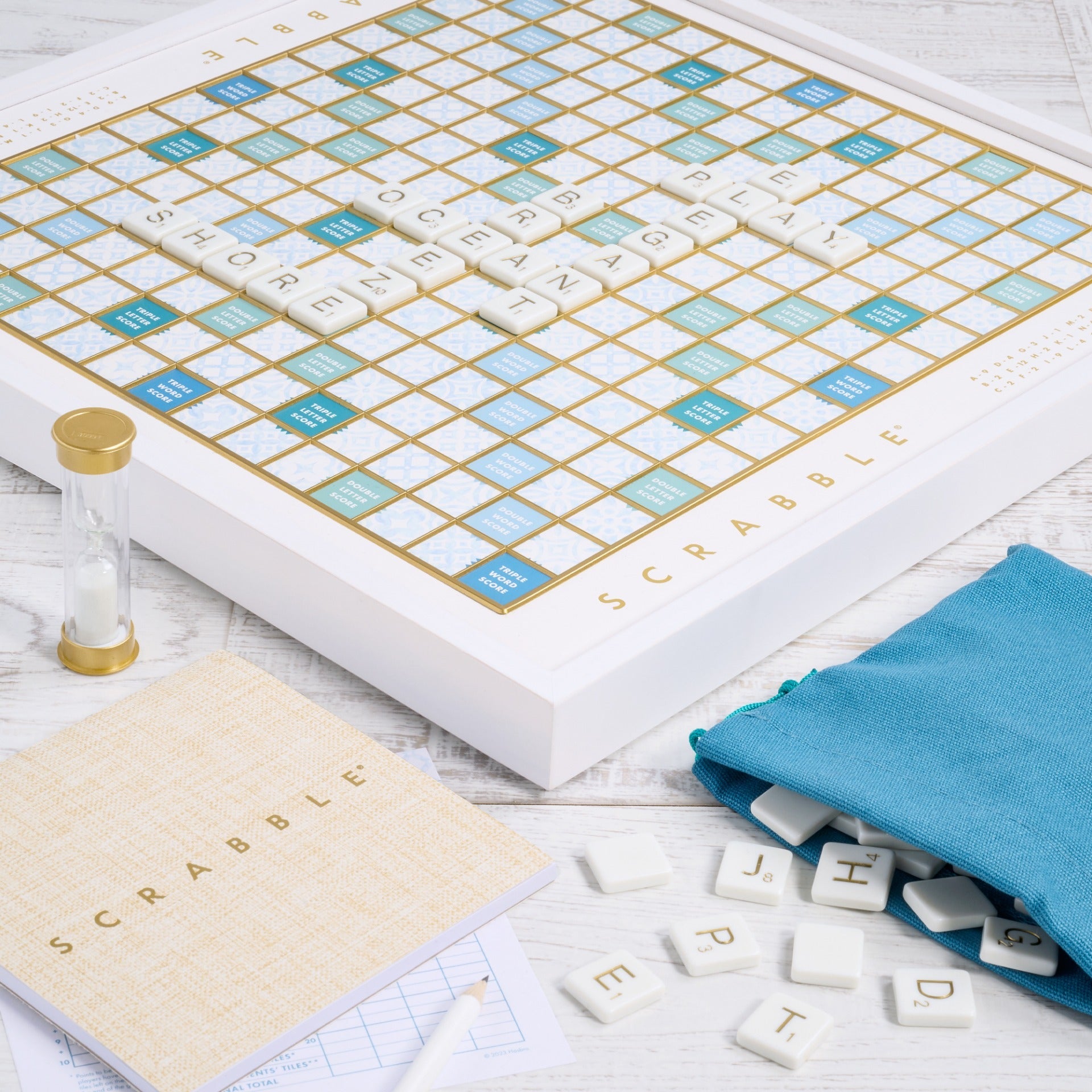 The Scrabble Bianco Edition by WS Game Company features a wood-framed board with letter tiles, a scorepad, pencil, sand timer, and a teal bag for extra tiles. Its ocean breeze design enhances the classic wooden look.