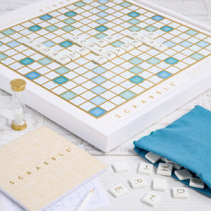 The Scrabble Bianco Edition by WS Game Company features a wood-framed board with letter tiles, a scorepad, pencil, sand timer, and a teal bag for extra tiles. Its ocean breeze design enhances the classic wooden look.