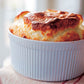 A golden-brown soufflé rises above the rim of a white, ribbed ceramic baking dish, sitting on a light-colored cloth—a classic touch inspired by recipes from Barefoot in Paris by Penguin Random House.