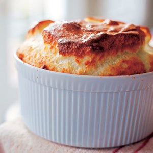 A golden-brown soufflé rises above the rim of a white, ribbed ceramic baking dish, sitting on a light-colored cloth—a classic touch inspired by recipes from Barefoot in Paris by Penguin Random House.