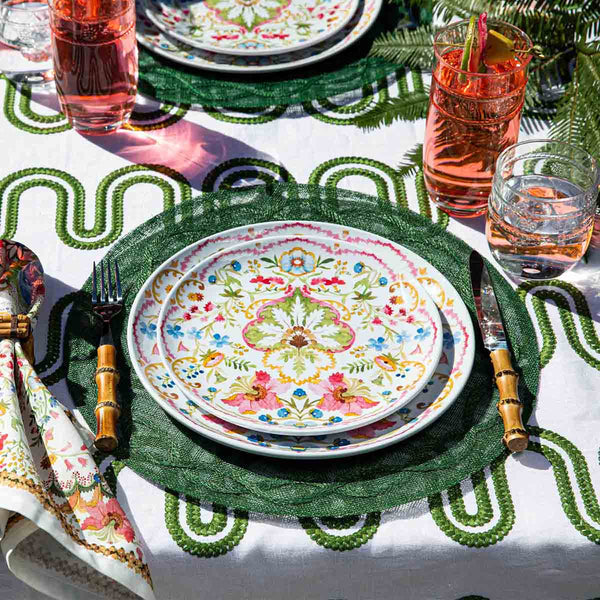 The Sofia Melamine Dinnerware Collection by Juliska, paired with green placemats, bamboo-handled cutlery, and a white tablecloth with a green wavy design, brings Fantasy garden charm to outdoor entertaining.