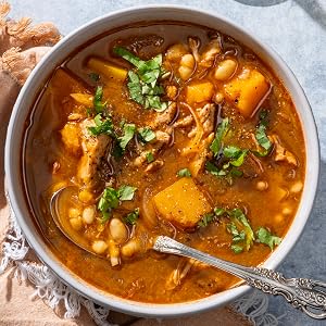 A bowl of hearty soup with beans, shredded meat, and chunks of squash, garnished with chopped herbs and a spoon resting inside—just one of the delicious high fiber and high protein recipes you’ll find in The Complete High-Protein Cookbook from Penguin Random House.