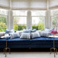 A blue velvet sofa with patterned blue-and-white pillows sits in a bright bay window alcove, flanked by small gold side tables—one holding pink flowers, the other featuring the Fia Table in aged brass by Made Goods. Trees are visible outside.