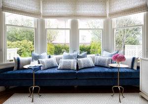 A blue velvet sofa with patterned blue-and-white pillows sits in a bright bay window alcove, flanked by small gold side tables—one holding pink flowers, the other featuring the Fia Table in aged brass by Made Goods. Trees are visible outside.