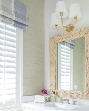 Bathroom vanity with the Made Goods Amani Rectangular Abaca Rope Mirror, brass light fixture with white shades, marble countertop, sink, soap dispenser, white towel, purple flower, and a window with crisp white shutters.
