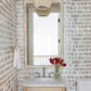 A small bathroom features patterned wallpaper, a marble-topped wood vanity, a Made Goods Pierson Mirror in Polished Bone above the sink, a white towel, toilet, and a vase of red flowers.