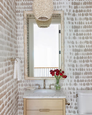 A small bathroom features patterned wallpaper, a marble-topped wood vanity, a Made Goods Pierson Mirror in Polished Bone above the sink, a white towel, toilet, and a vase of red flowers.