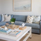 A light blue sofa with patterned pillows and textured upholstery sits behind the Made Goods Jarin Coffee Table in Designer White, which holds books, a candle, and decor; a large beach photo hangs on the wall.