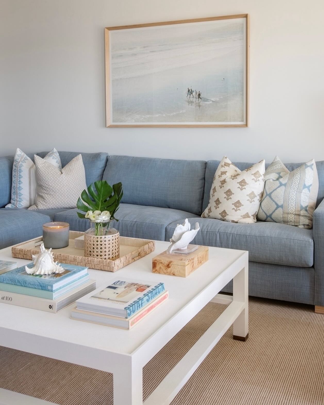 A light blue sofa with patterned pillows and textured upholstery sits behind the Made Goods Jarin Coffee Table in Designer White, which holds books, a candle, and decor; a large beach photo hangs on the wall.