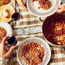 Three people serve tomato risotto from a pot onto plates at a checkered table with wine glasses and a plate of cheese and bread, capturing the inviting atmosphere of sharing authentic Italian food inspired by cherished recipes from Pasta Grannies: Comfort Cooking by Chronicle Books.