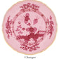 The Oriente Italiano Porpora Dinnerware Collection by Ginori 1735 features a pink charger plate with scalloped edges, gold trim, and an ornate floral and landscape motif inspired by Italian elegance.