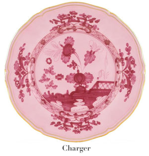 The Oriente Italiano Porpora Dinnerware Collection by Ginori 1735 features a pink charger plate with scalloped edges, gold trim, and an ornate floral and landscape motif inspired by Italian elegance.