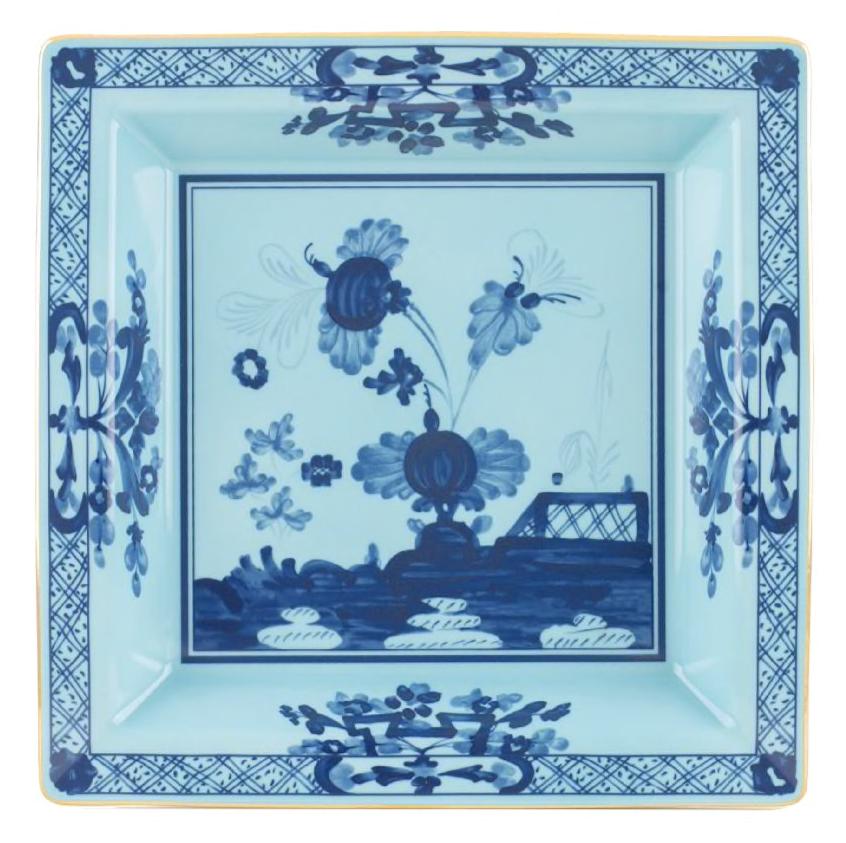 The Oriente Italiano Iris Tray Collection by Ginori 1735 features square porcelain plates in the Antico Doccia shape, decorated with blue floral and landscape patterns on a light blue background and classic iris blue-inspired borders.