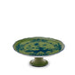 The Oriente Italiano Malachite cake stand by Ginori 1735 features a green ceramic body with a blue exotic floral pattern, scalloped edge, and pedestal base, shown against a plain white background.