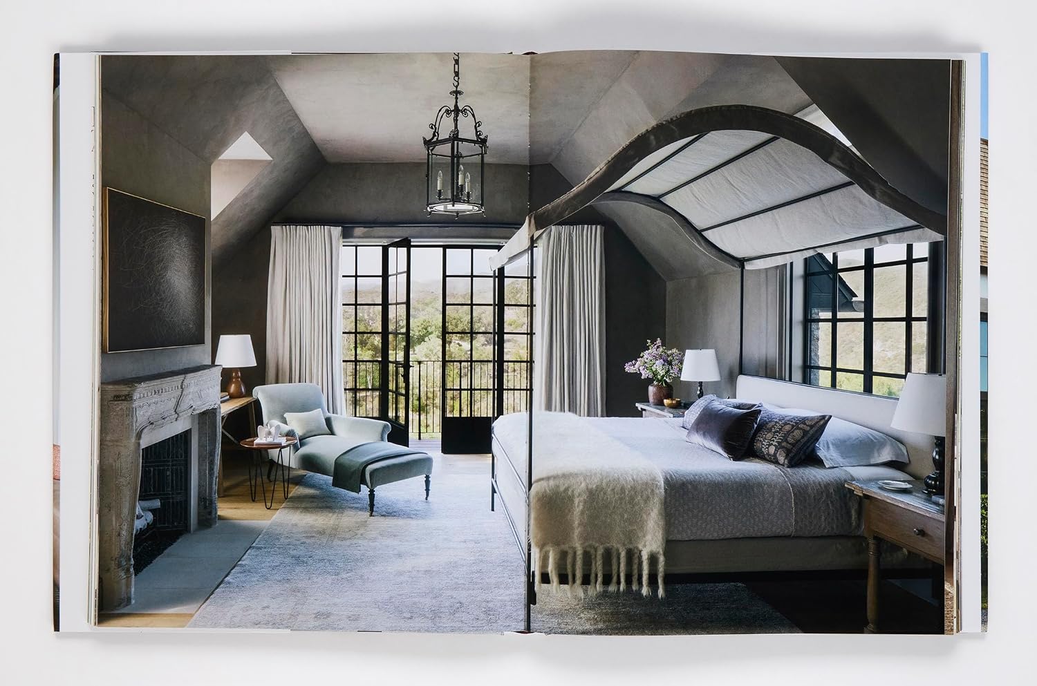 A modern bedroom inspired by Ray Booth: The Expressive Home from Rizzoli features a spacious bed, cozy fireplace, elegant lounge chair, and expansive floor-to-ceiling windows that invite abundant natural light. Decorated in neutral tones with contemporary furnishings, the space achieves a timeless aesthetic ideal for sophisticated homes.
