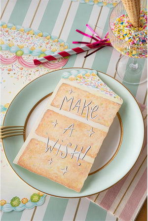 The Hester & Cook Cake Slice Table Accent, shaped like a birthday cake slice with "MAKE A WISH!", serves as charming table decor beside a glass holding an ice cream cone with sprinkles and colorful straws.
