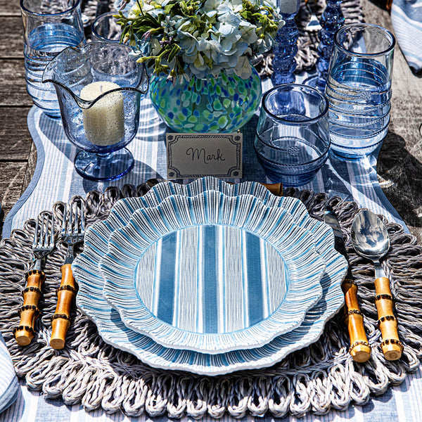 A table setting with blue and white striped plates, bamboo-handled cutlery, blue glassware, a floral centerpiece, Juliska Straw Loop Placemat Collection for natural texture, and a place card reading "Mark.