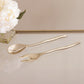 The Beatriz Ball Sierra Modern Maia Salad Servers, in radiant gold, are displayed side by side on a reflective surface beside a vase of white flowers.