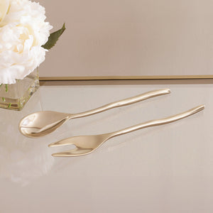 The Beatriz Ball Sierra Modern Maia Salad Servers, in radiant gold, are displayed side by side on a reflective surface beside a vase of white flowers.
