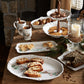 A table set with mugs of tea, cookies, cakes, and pastries on Juliska’s Berry & Thread North Pole Dinnerware and Serveware Collection, featuring winter scenes, beside a large candle in a glass hurricane.