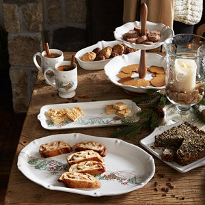 A table set with mugs of tea, cookies, cakes, and pastries on Juliska’s Berry & Thread North Pole Dinnerware and Serveware Collection, featuring winter scenes, beside a large candle in a glass hurricane.