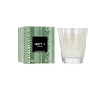 The Nest Birchwood Pine Collection candle, in clear patterned glass beside its green box with a black-and-white label, fills your space with a winter forest scent for a festive atmosphere.